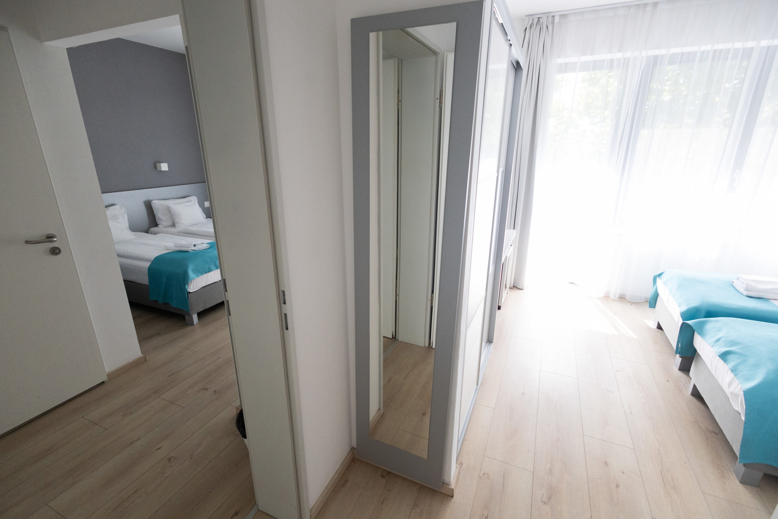 A képen egy összenyitható családi szoba látható. A felvétel a közös ajtóból készült, így mindkét helyiség belátható – jól kivehető, ahogy a fehér ágyneműt türkizkék ágysál díszíti.
