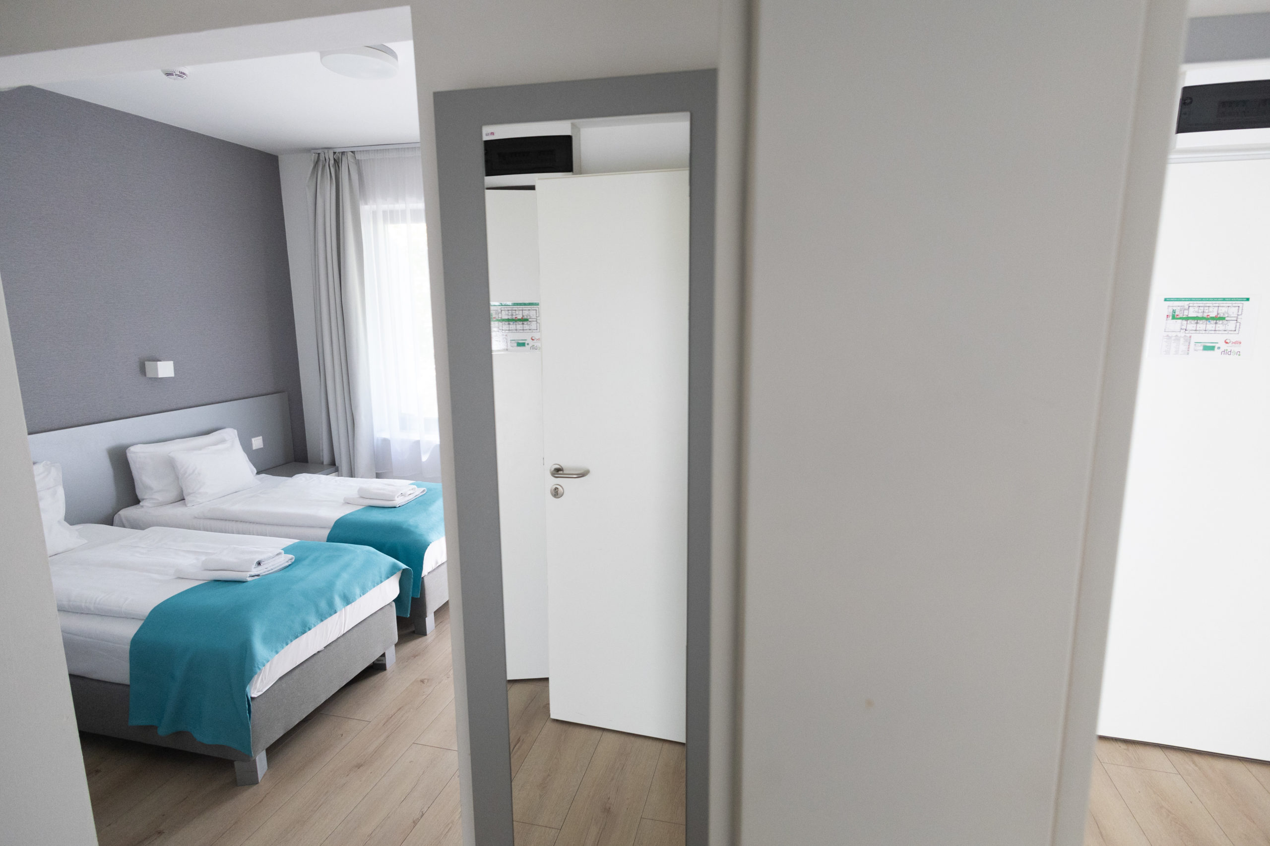 A képen egy összenyitható családi szoba látható. A felvétel a közös ajtóból készült, így mindkét helyiség belátható – jól kivehető, ahogy a fehér ágyneműt türkizkék ágysál díszíti.
