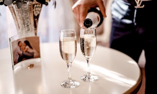 A képen egy asztal látható, amin van két pezsgős pohár és épp a felszolgáló tölti a pezsgőt a poharakba.